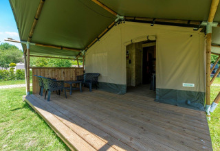 Safari-Zelt mit überdachter Holzterrasse, Stühlen und Tisch, umgeben von grüner Natur im Freien.
