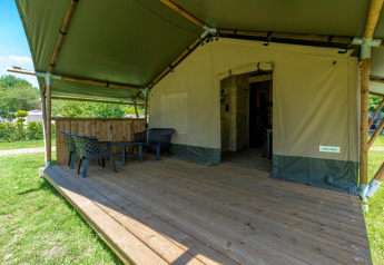 Tenda safari con veranda in legno coperta, sedie e tavolino, immersa nel verde della natura.