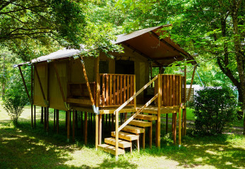 Tienda safari llamada Safaritent 4 para 4 personas, elevada sobre pilotes y rodeada de árboles verdes.