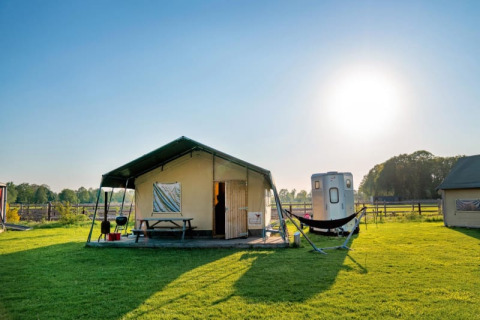 Safari-Zelt Luxe Ranchtent auf einer sonnigen Wiese mit Hängematte, Holztisch und Grill im Freien.