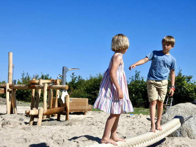 Dos niños juegan descalzos en un parque de arena con estructuras de madera en un alojamiento glamping.