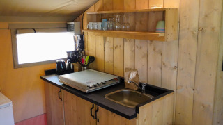 Kleine kitchenette in een safaritent met spoelbak, kookplaat, keukengerei en open planken met glazen.