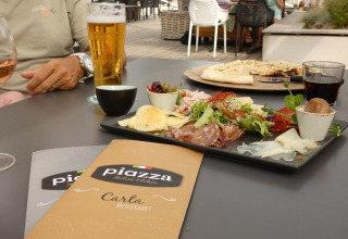 Comida al aire libre en glamping con antipasto italiano, bebidas y cartas sobre la mesa en ambiente relajado.