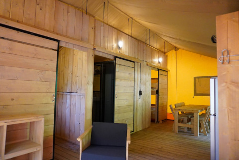 Intérieur d'une tente safari avec murs en bois, coin repas, frigo et chaises au Camping De Rammelbeek.