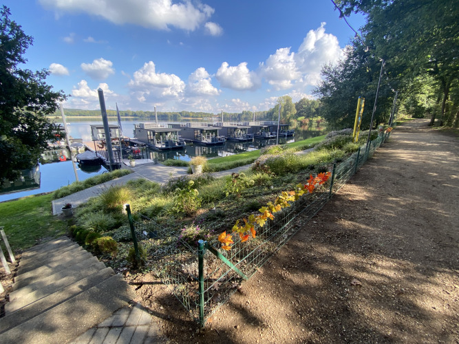 Blick auf Hausboot-Marina Mookerplas in den Niederlanden mit Hausbooten, Blumen und einem Weg am Wasser.