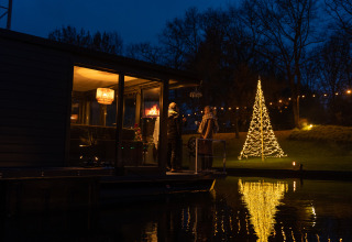 Avondbeeld van een houseboat bij Marina Mookerplas met verlichte kerstboom en mensen op het terras.
