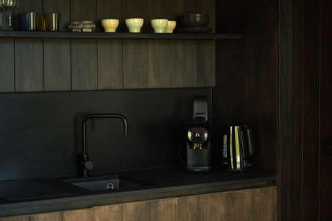 Pequeña cocina oscura en Nokken Forest Cabin con fregadero negro, cafetera, hervidor y tazas.