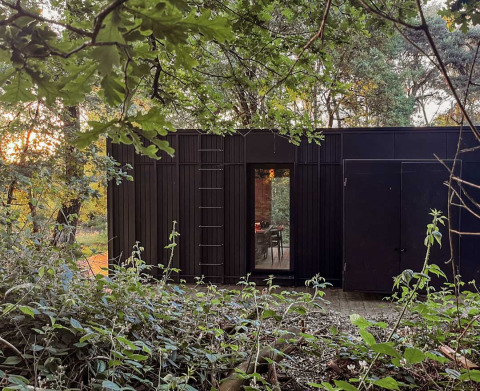 Schwarze Nokken Forest Cabin im Wald bei Wilsumer Berge, Deutschland, umgeben von grüner Natur.