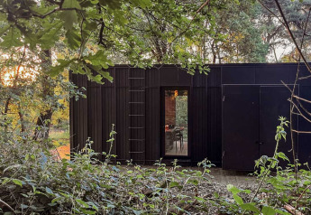 Black Nokken Forest Cabin in the woods at Wilsumer Berge, Germany, surrounded by lush green trees.