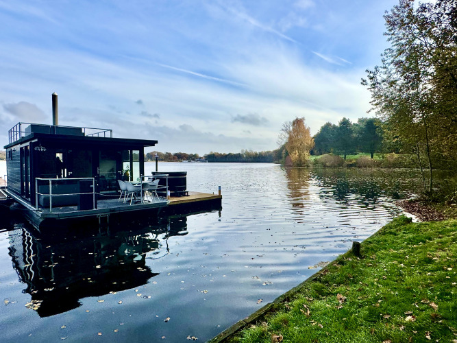 Hausboot mit Dachterrasse und Whirlpool auf ruhigem See, umgeben von Bäumen, bei Marina Mookerplas, Niederlande.