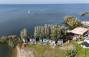 Luftfoto af små hytter ved søbredden i Marina Parcs Almere, Holland, omgivet af grønne træer og vand.