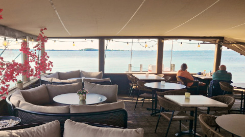 Gezellige overdekte terras met comfortabele banken en uitzicht op het water, twee mensen genieten ervan.
