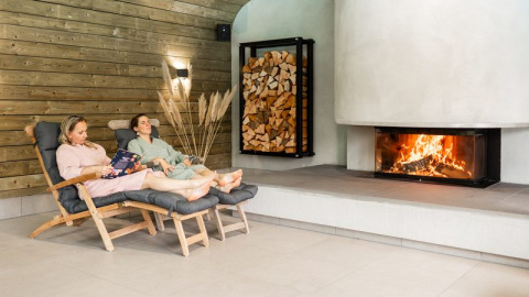 Two people relax on lounge chairs by a fireplace in a cozy glamping accommodation with wood decor.