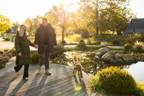 Couple holding hands walks with their dog by a pond at a scenic glamping site in the golden sunlight.