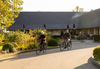 To personer cykler foran en glampingbolig omgivet af grønne planter og sollys ved en moderne hytte.
