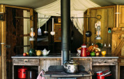 Rustic kitchen inside a safari tent with wood cabinets, vegetables, and kettle, cozy Dutch glamping style.