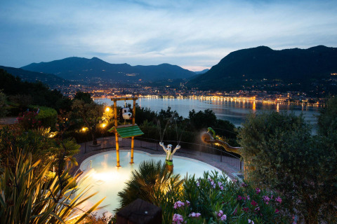 Abendaufnahme vom Weekend Glamping Resort mit Kinderpool und Blick auf den beleuchteten See in der Lombardei.