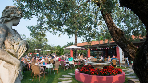 Udendørs restaurantområde med gæster, springvand, statue og træer på Weekend Glamping Resort i Lombardiet.
