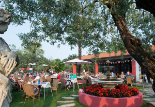 Outdoor dining area with guests, a fountain, statue, and trees at Weekend Glamping Resort in Lombardy, Italy.