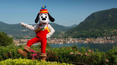 Maskottchen im roten Overall posiert vor See und Bergen bei Weekend Glamping Resort in der Lombardei.