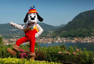 Maskot i rød dragt poserer foran sø og bjerge ved Weekend Glamping Resort i Lombardiet, Italien.