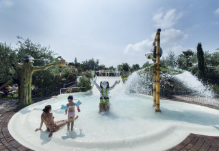Children and an adult play in a splash pool with water features and statues at Weekend Glamping Resort, Lombardy.