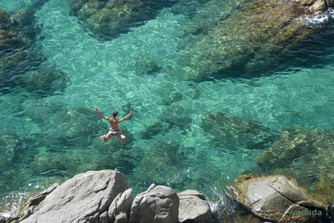 Persona saltando desde rocas al agua cristalina en Wecamp L'Escala Punta Mila, Cataluña, España.