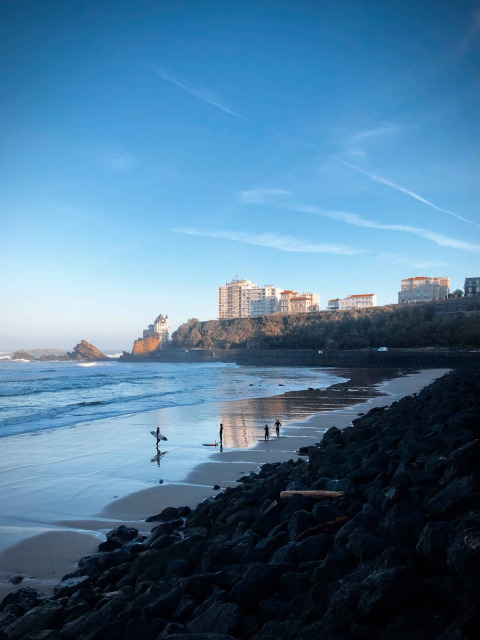 Surfisti su una spiaggia vicino a San Sebastián nei Paesi Baschi, Spagna, con rocce e edifici sullo sfondo.