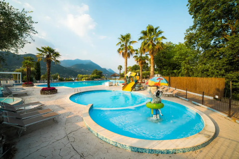 Poollandschaft mit Palmen und Wasserrutsche im Weekend Glamping Resort in der Lombardei, Italien, mit Bergen im Hintergrund.
