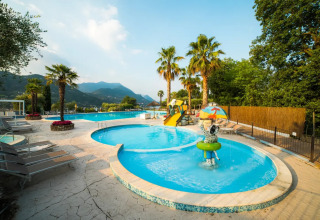 Poolområde med palmer og rutsjebane på Weekend Glamping Resort i Lombardiet, Italien, med bjerge i baggrunden.
