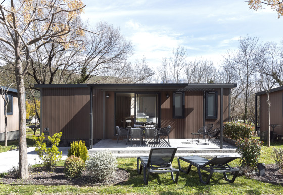 Mobile home FAMILY XL-lodge med udendørs terrasse, stole og solsenge i grøn og frodig have.