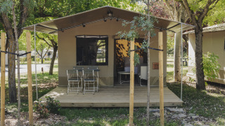 Cabaña tipo safari con terraza de madera, mesa y sillas al frente, rodeada de árboles verdes.