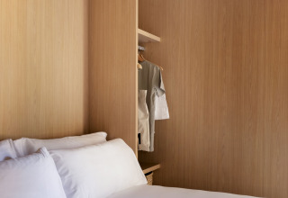 Glampingkamer met houten wanden, fris wit beddengoed en een open kledingkast met enkele hemden.