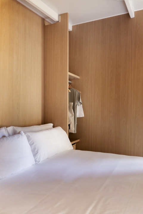 Acogedora habitación de glamping con cama cómoda, paneles de madera clara y ropa colgada en perchero.