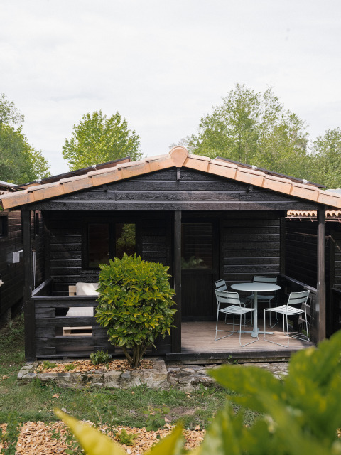Pequeña casa negra de madera con terraza y muebles de exterior en Wecamp Cudillero, España, rodeada de vegetación.