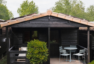 Pequeña casa negra de madera con terraza y muebles de exterior en Wecamp Cudillero, España, rodeada de vegetación.