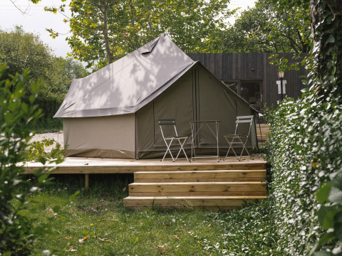 Tenda safari su pedana in legno con due sedie e un tavolino, immersa nel verde tra alberi e natura.