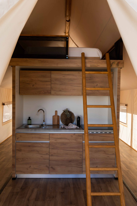 Interieur van een safaritent met een moderne kitchenette, houten ladder en slaapzolder bovenin.