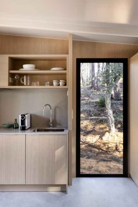 Coin cuisine moderne pour glamping avec armoires en bois clair et porte vitrée donnant sur la forêt.