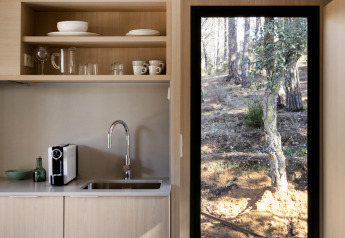 Coin cuisine moderne pour glamping avec armoires en bois clair et porte vitrée donnant sur la forêt.