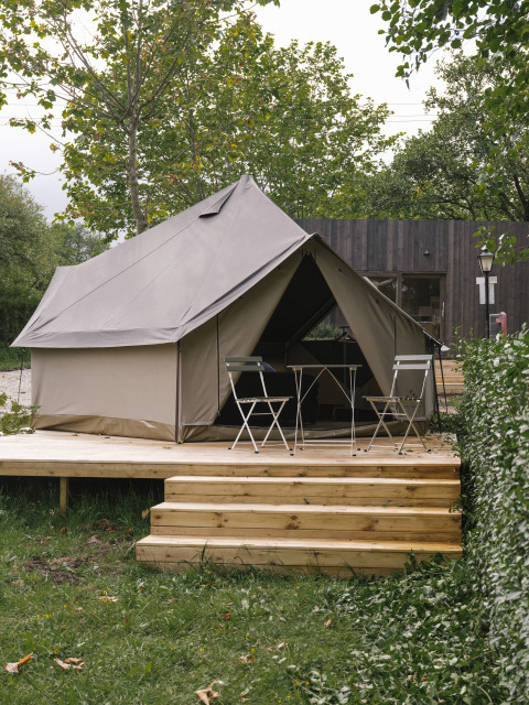 Safari-Zelt auf einer Holzterrasse mit Klappstühlen und Tisch, umgeben von grünen Bäumen und Sträuchern.