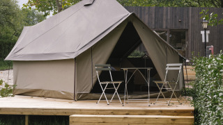 Safari-Zelt auf einer Holzterrasse mit Klappstühlen und Tisch, umgeben von grünen Bäumen und Sträuchern.