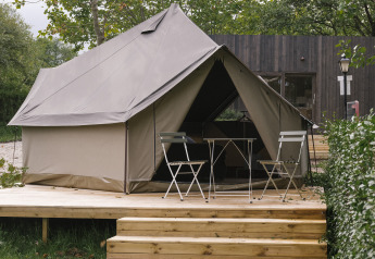 Safari-Zelt auf einer Holzterrasse mit Klappstühlen und Tisch, umgeben von grünen Bäumen und Sträuchern.