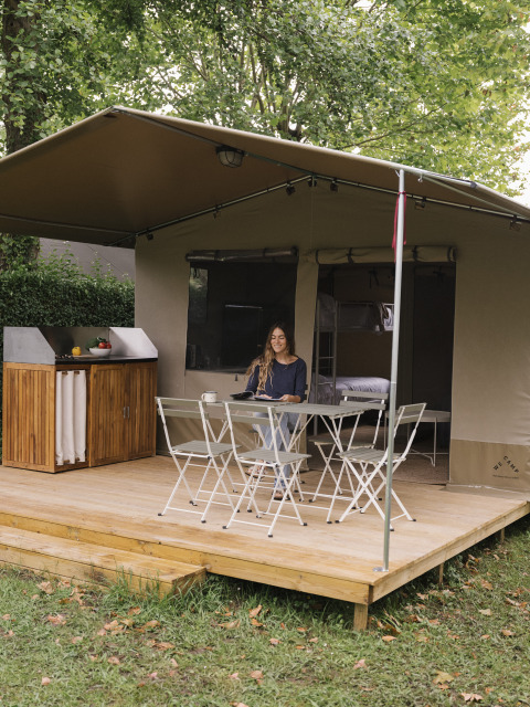 Une femme est assise à une table sur la terrasse d'une tente de glamping entourée d'arbres et de verdure.