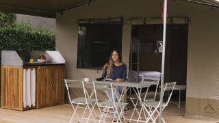 Eine Frau sitzt an einem Tisch auf der Terrasse eines Glamping-Zelts, umgeben von grüner Natur und Bäumen.