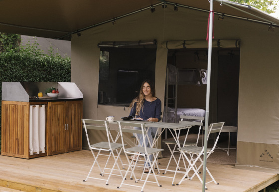 Une femme est assise à une table sur la terrasse d'une tente de glamping entourée d'arbres et de verdure.