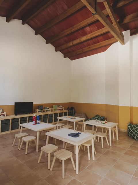 Sala infantil luminosa con mesas, taburetes, juguetes, TV y techo de vigas en Wecamp Jávea, Valencia.