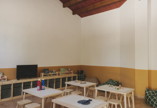 Bright playroom with small tables, stools, toys, TV, and wooden beam ceiling at Wecamp Jávea, Valencia.