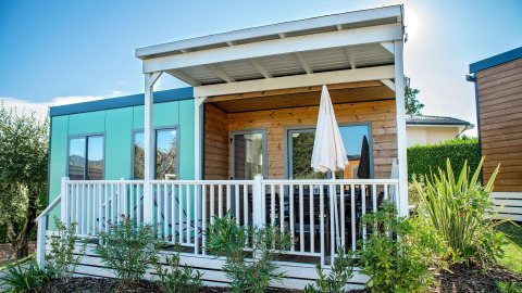 Exterior view of the Next L Deluxe lodge with covered porch, white railing, and lush greenery in front.