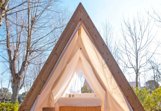 A-frame wooden cabin with bed and curtains in sunlight at Wecamp Santa Cristina, Catalonia, Spain.